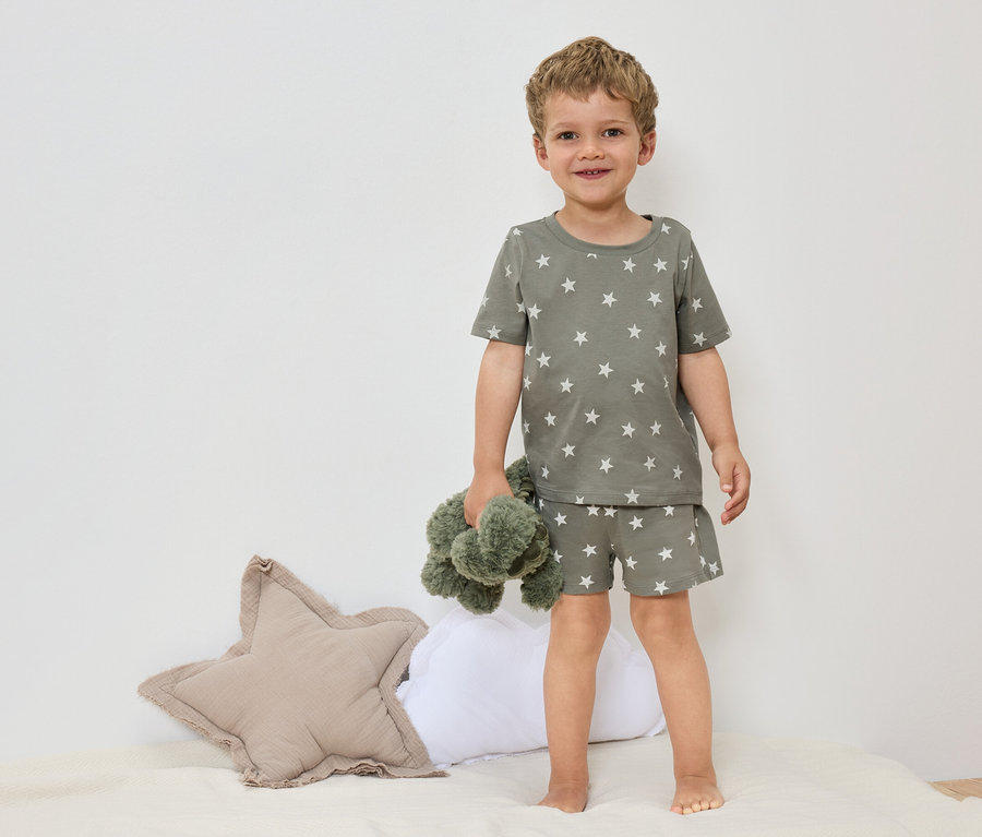 Malý chlapec stojí bosý na posteli v olivovozelenom pyžame s hviezdami a drží zelenú plyšovú hračku.