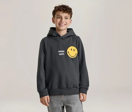 Usmiaty chlapec v tmavosivej mikine s kapucňou so žltou smajlíkovou tvárou, ktorá má namiesto oka X a efekt topenia, a nápisom „CHOOSE HAPPY“. Má na sebe svetlosivé džínsy.