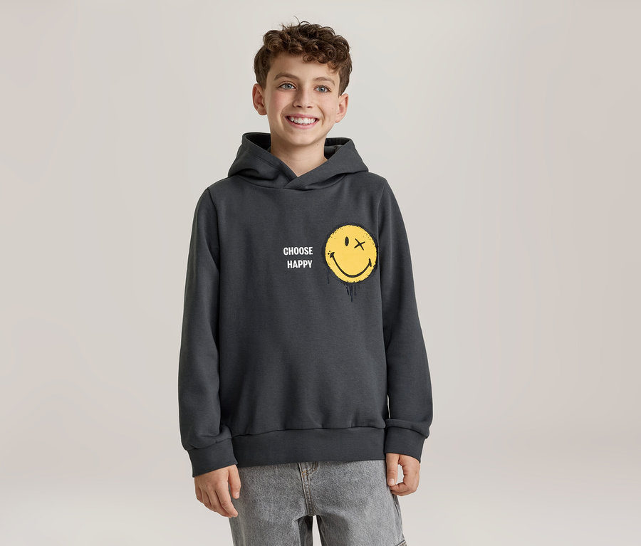 Usmiaty chlapec v tmavosivej mikine s kapucňou so žltou smajlíkovou tvárou, ktorá má namiesto oka X a efekt topenia, a nápisom „CHOOSE HAPPY“. Má na sebe svetlosivé džínsy.