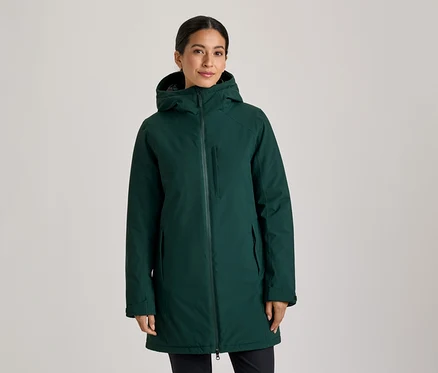 Žena stojí v tmavozelenom kabáte s kapucňou, zapnutom na zips, s rukami vo vreckách a pozerá do kamery.