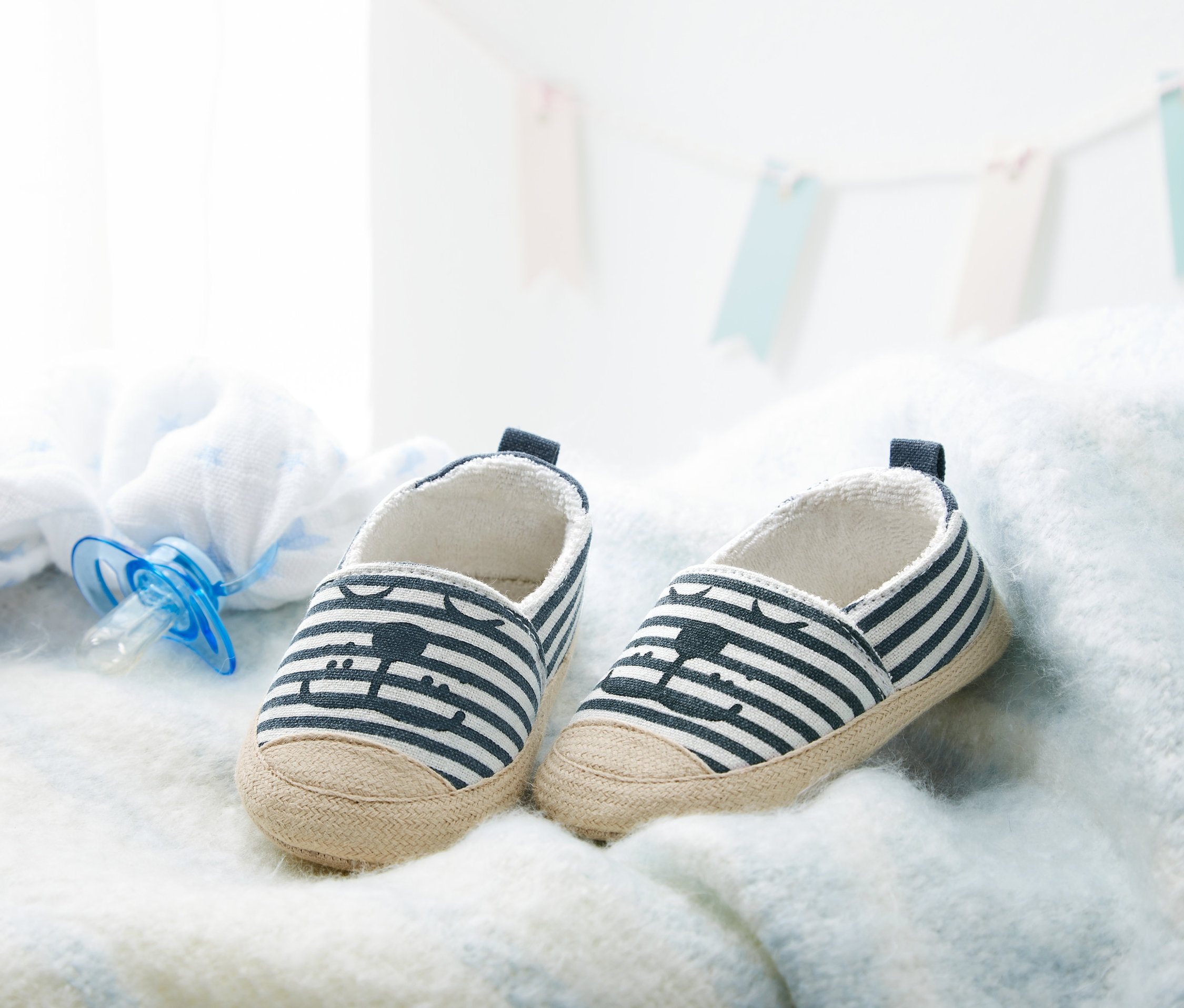 Dvoje topánky pre bábätká s modro-bielymi pruhmi ležia na bielej deke vedľa modrého cumlíka.