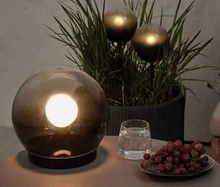 Na stolíku stojí guľové svietidlo z dymového skla, pohár vody a hrozno. V pozadí sú v kvetináči 2 guľové LED svietidlá.