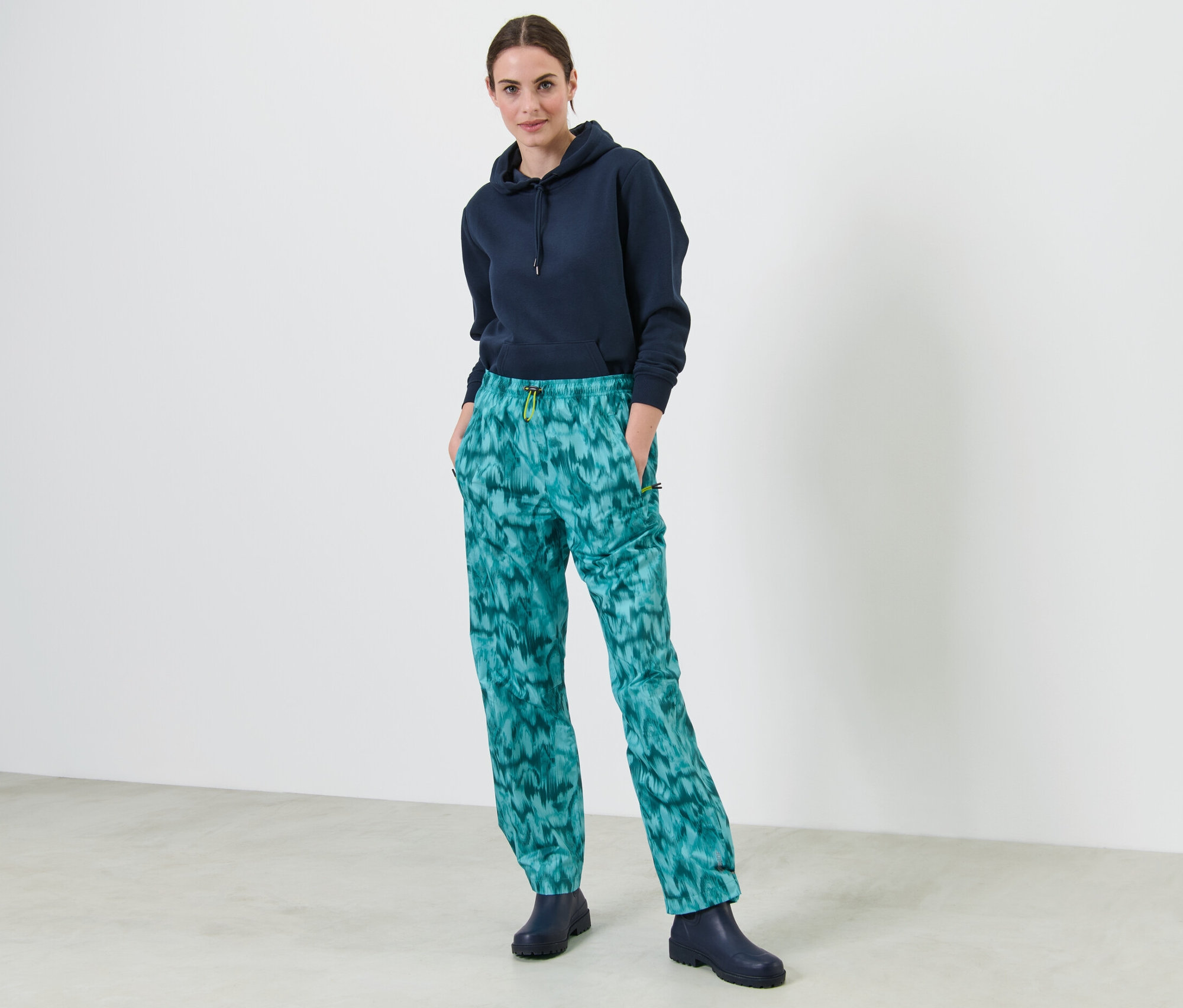 Žena stojí a má ruky vo vreckách nohavíc do dažďa s celoplošnou potlačou.