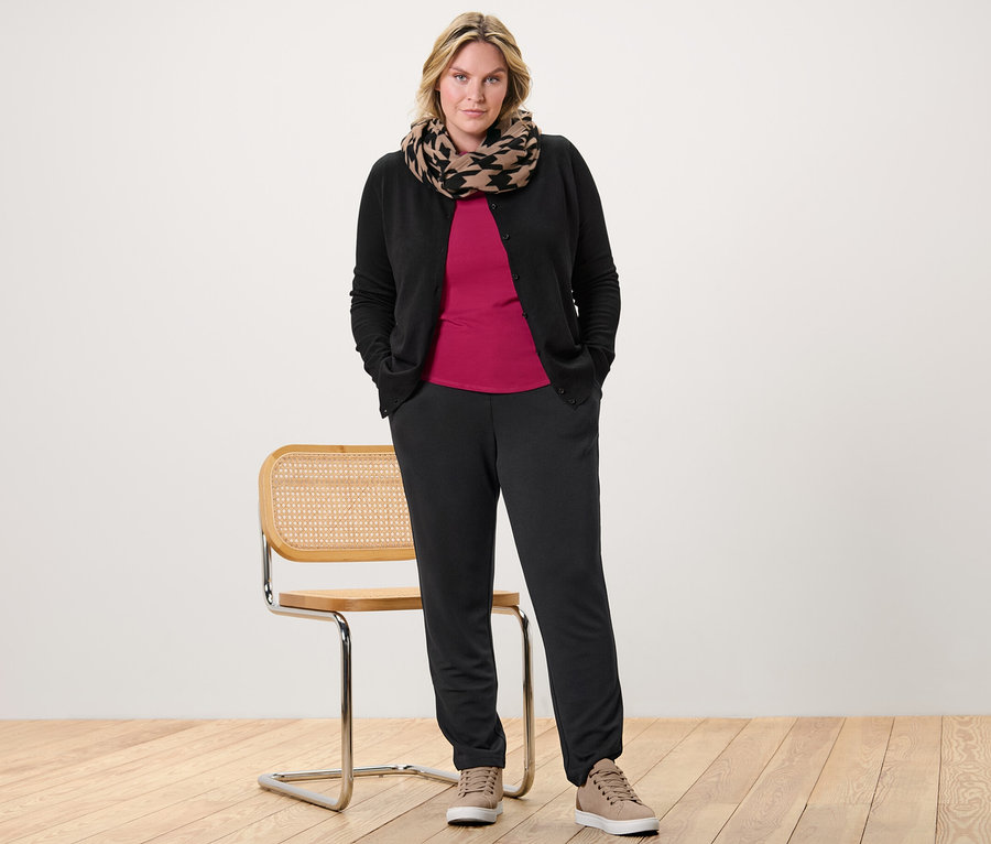 Žena pózuje v malinovo červenom tričku s trojštvrťovým rukávom, kardigáne z jemného úpletu a teplákoch.