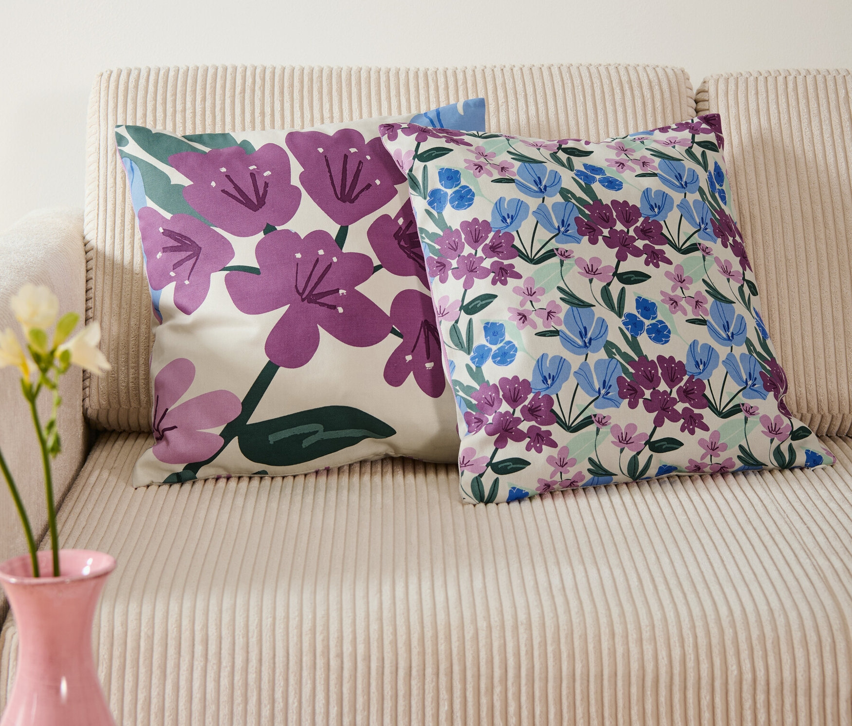 Dve obliečky na dekoračné účely s kvetinovými vzormi ležia na béžovej pohovke, nad nimi visí modrý obrázok s kvetmi vo váze.