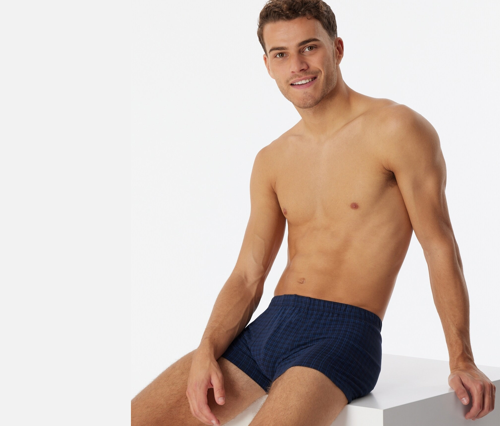 Muž sedí na bielej kocke a má na sebe kárované boxerky SCHIESSER.