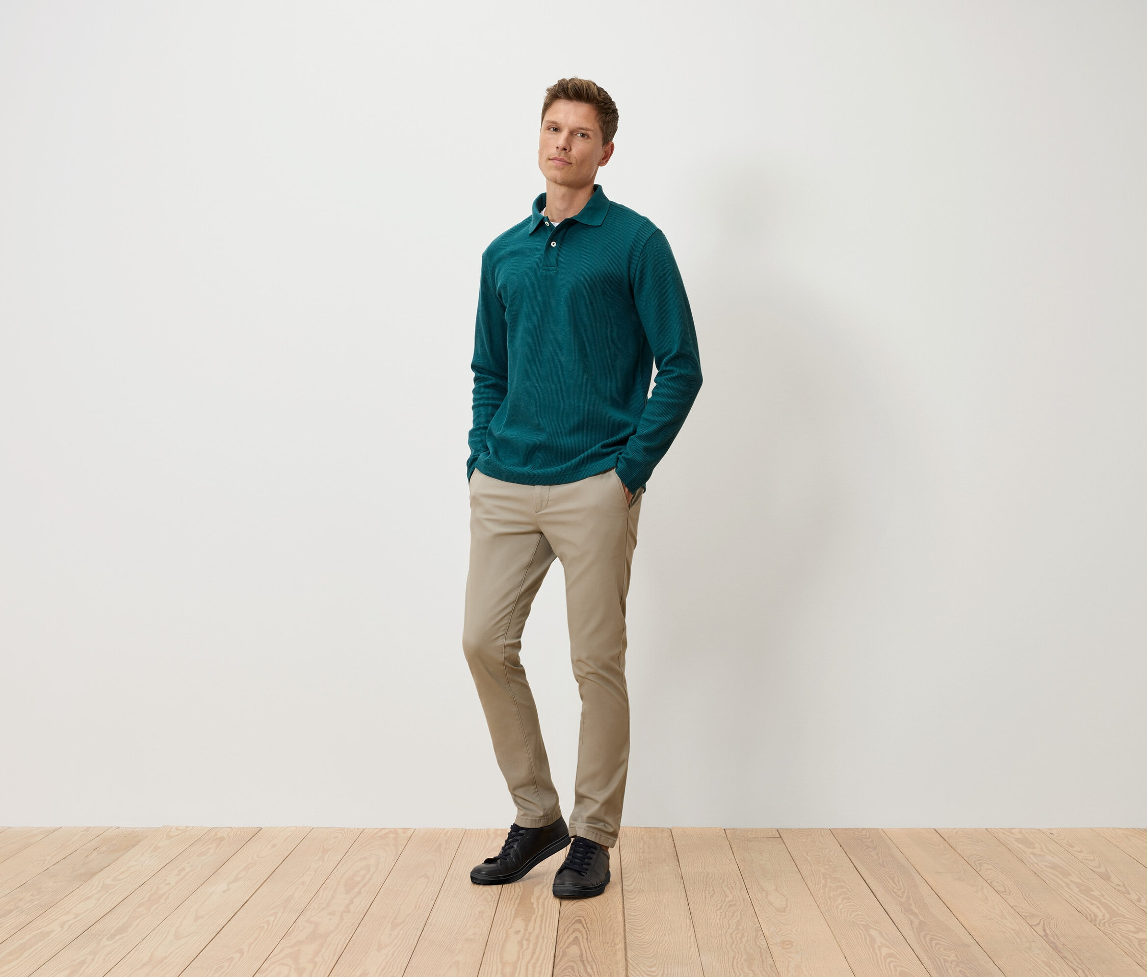 Muž stojí s rukami vo vreckách a má na sebe zelené polo tričko s dlhým rukávom, béžové nohavice a čierne topánky.