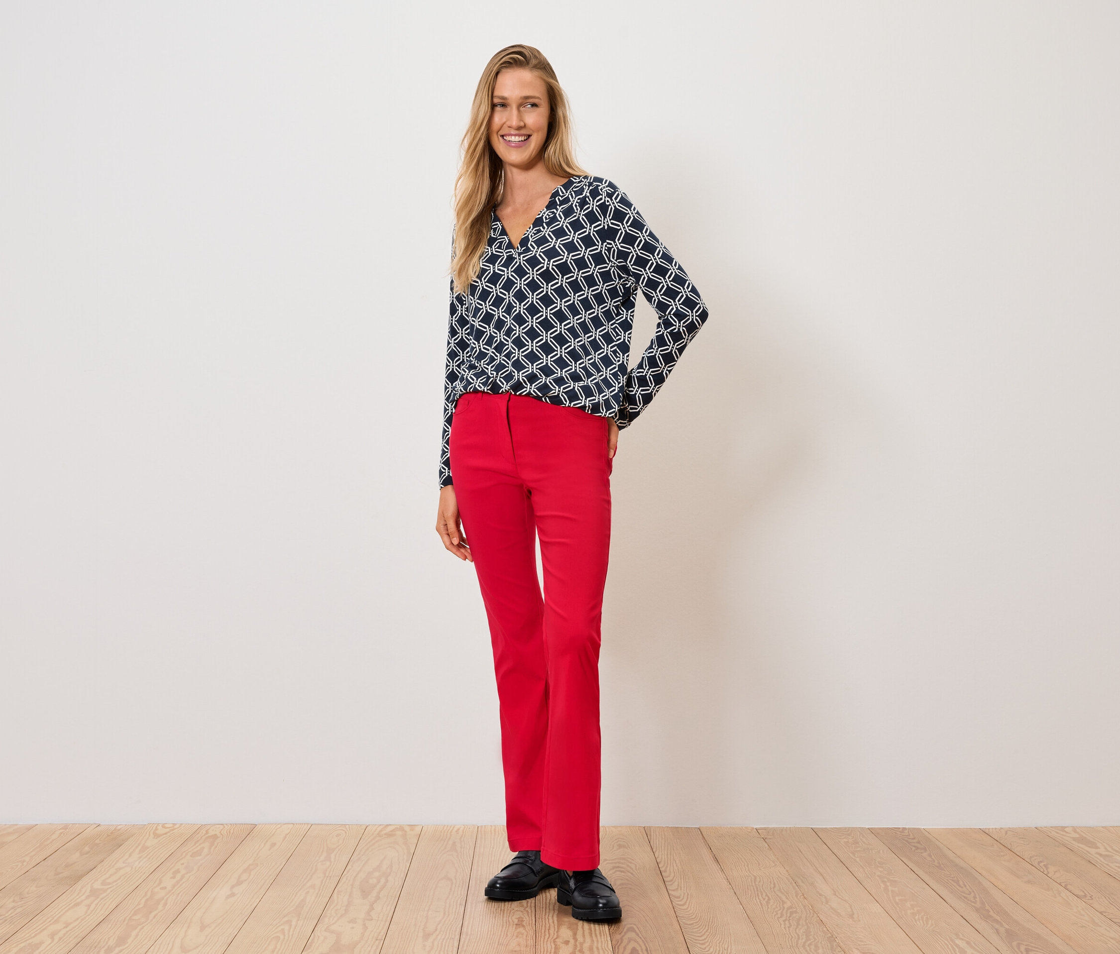 Žena v modro-bielom tope a červených nohaviciach stojí na drevenej podlahe.