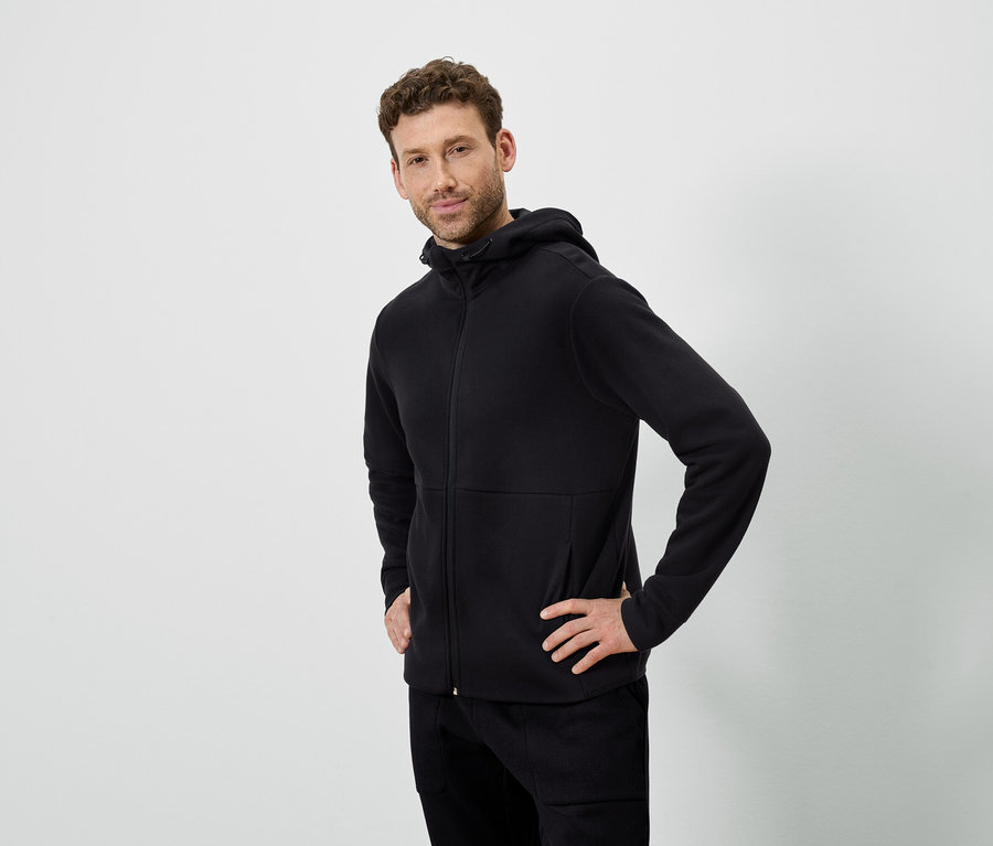 Muž pózuje na bielom pozadí. Má na sebe čiernu mikinu s kapucňou a čierne nohavice.