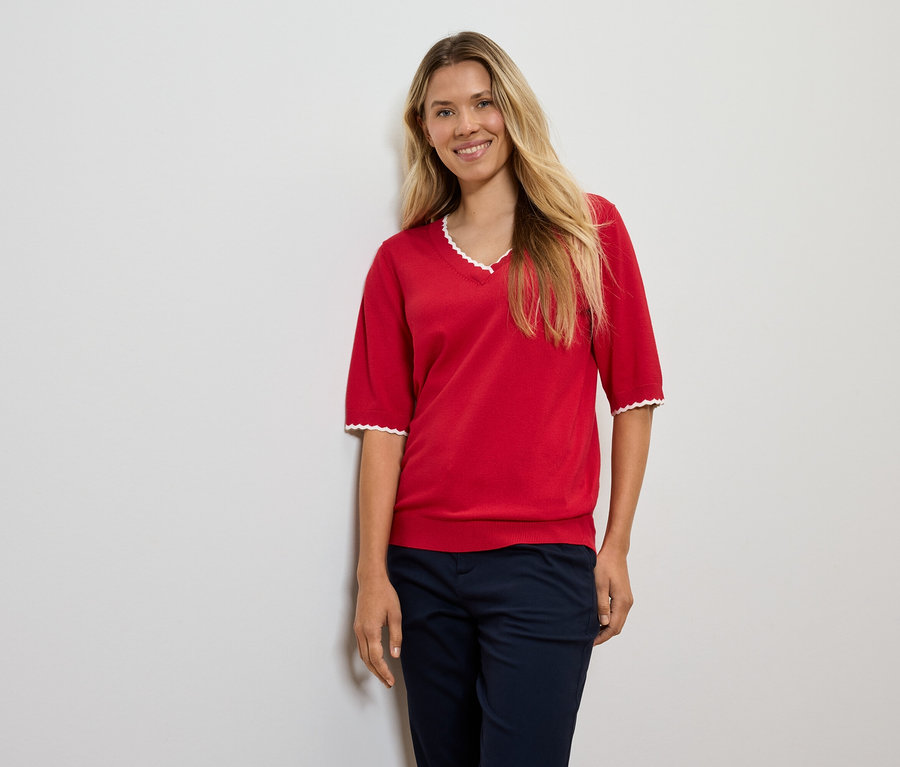 Žena s blond vlasmi pózuje v červenom svetri a tmavomodrých nohaviciach.