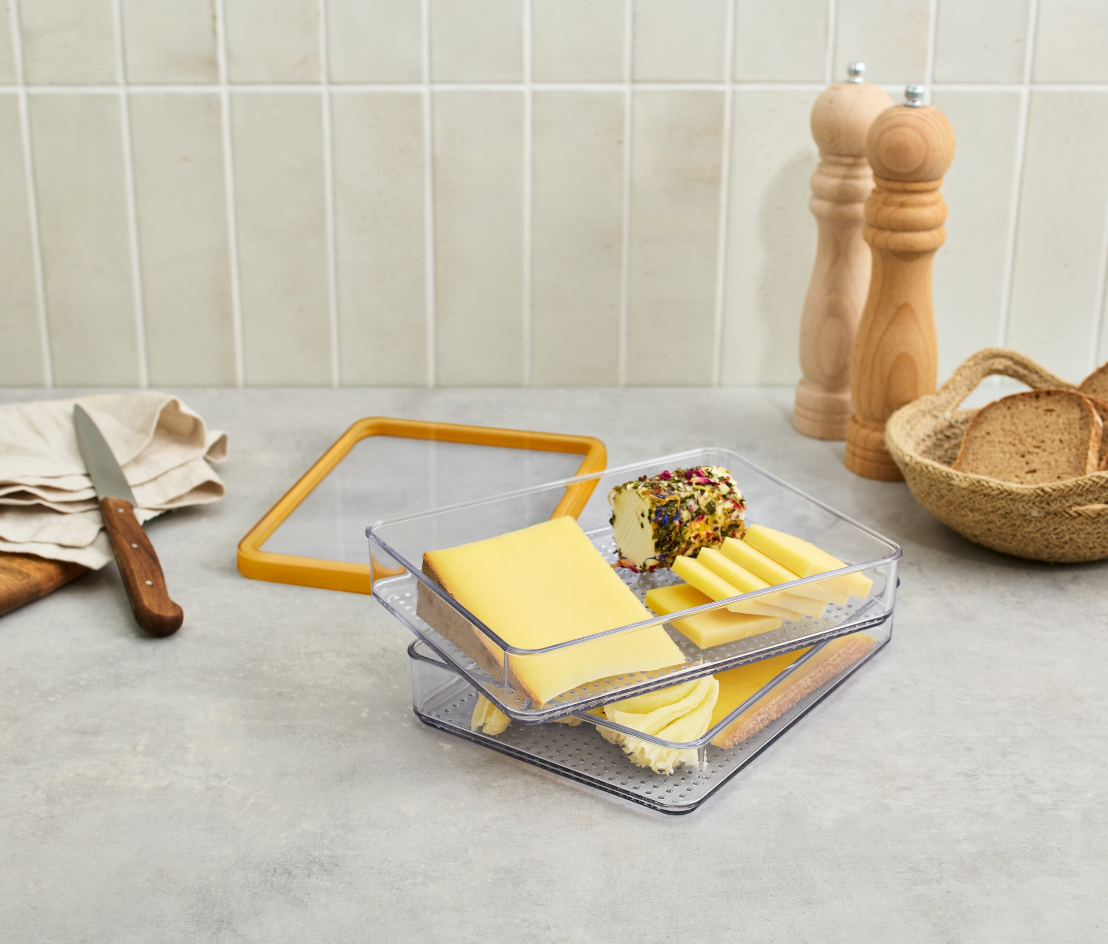 Na stole je syr v priehľadnej nádobe, hrozno, nôž, chlieb v košíku a mlynčeky na korenie.