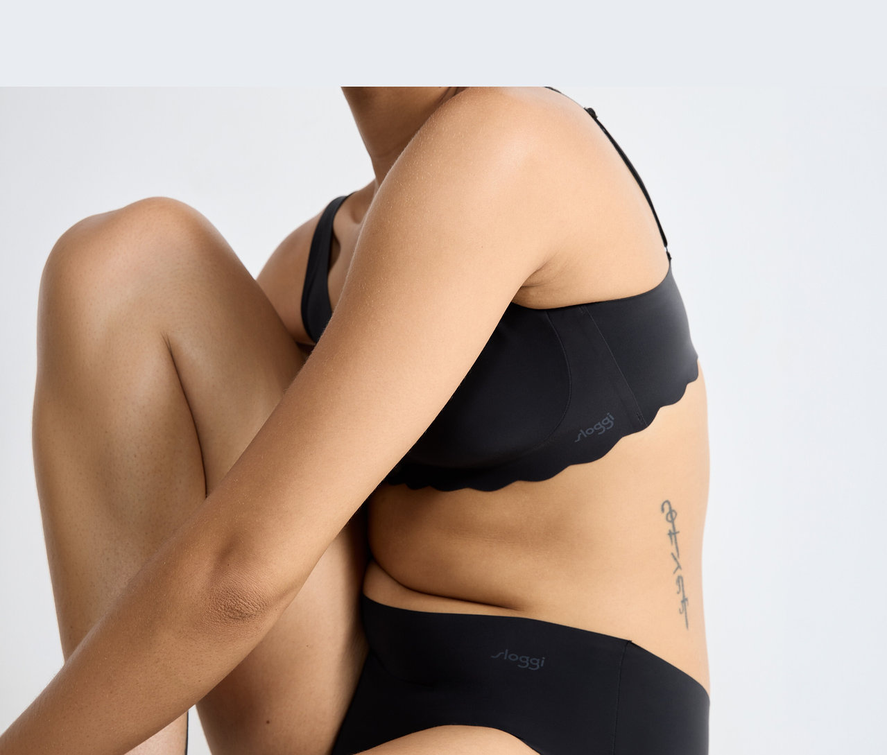 Osoba v čiernej podprsenke sloggi ZERO Microfibre 2.0 Soft bra s vlnitým okrajom a zodpovedajúcich čiernych nohavičkách sloggi, sediaca so zohnutými kolenami a prekríženými rukami. Na boku je viditeľné tetovanie.