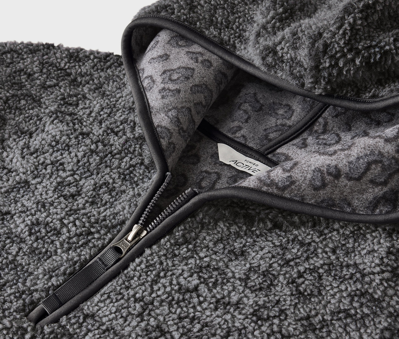 Detail šedej chlpaté mikiny s kapucňou so zipsom a šedým zvieracím vzorom na vnútornej strane kapucne.