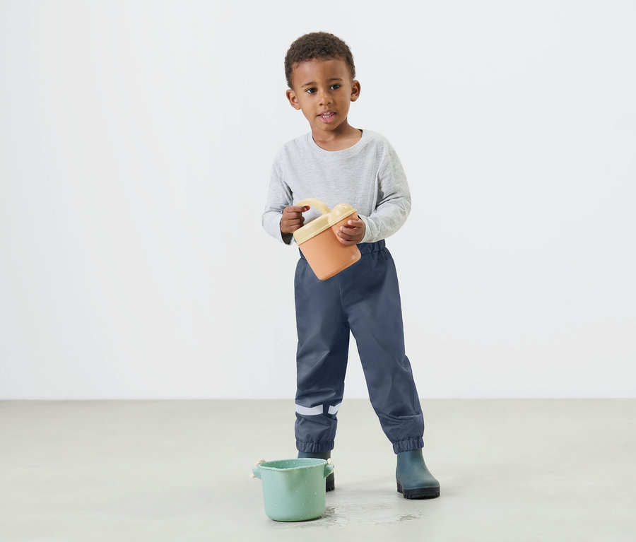 Malý chlapec drží v rukách oranžové vedro a má na sebe modré nepremokavé nohavice a gumáky.