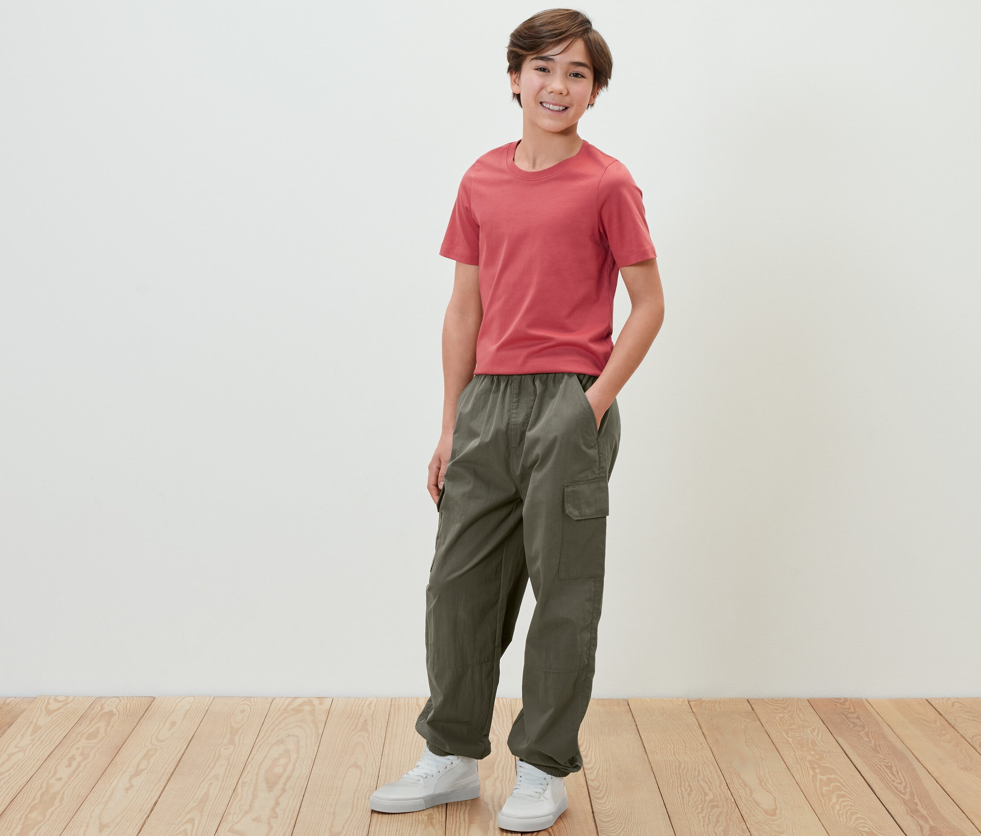 Chlapec v červenom tričku a detských cargo nohaviciach stojí pred bielou stenou.