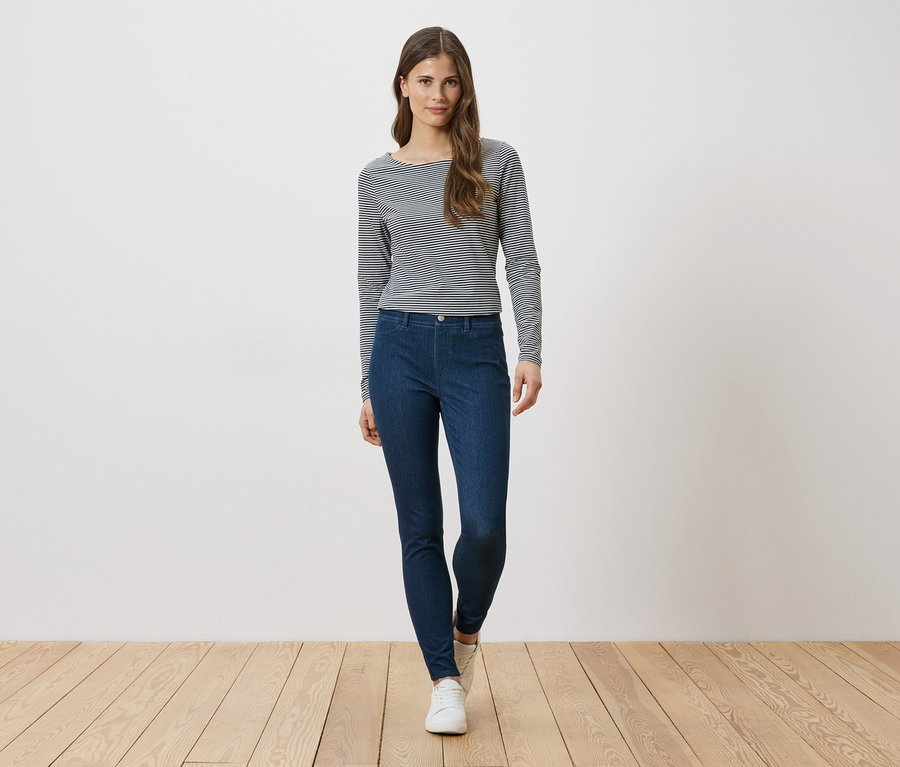 Žena v pruhovanom tričku s dlhým rukávom a modrých džínsoch stojí pred bielou stenou.