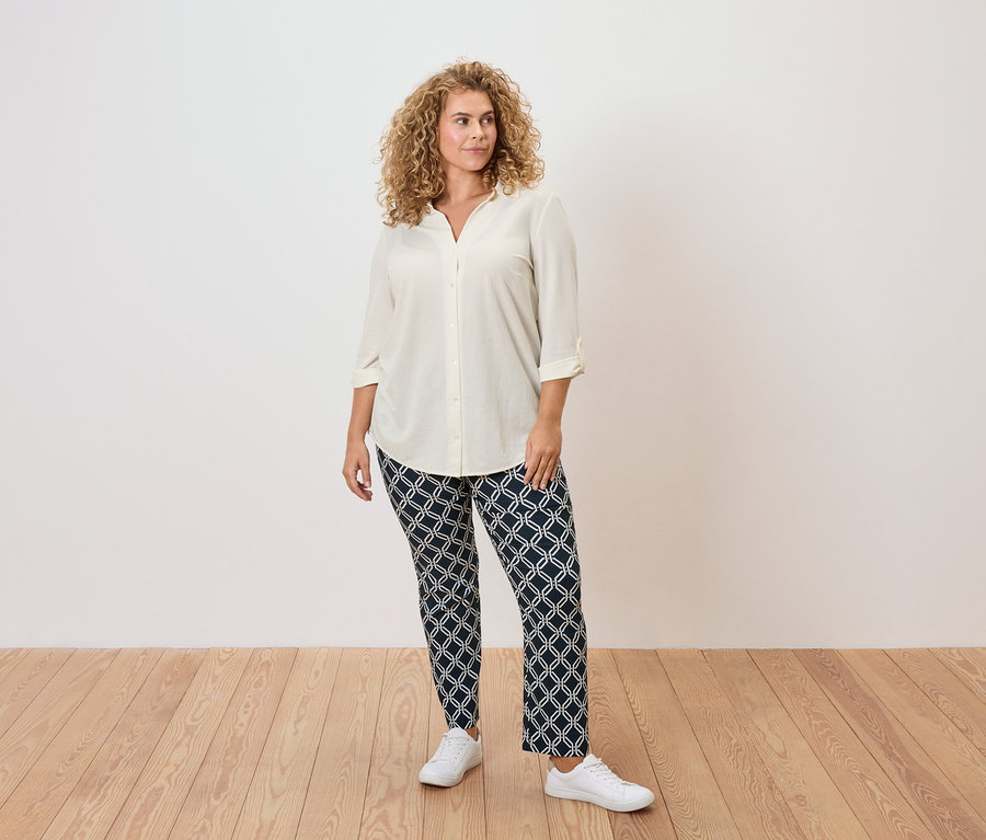 Žena s kučeravými vlasmi má na sebe krémovú blúzku, vzorované nohavice a biele tenisky.