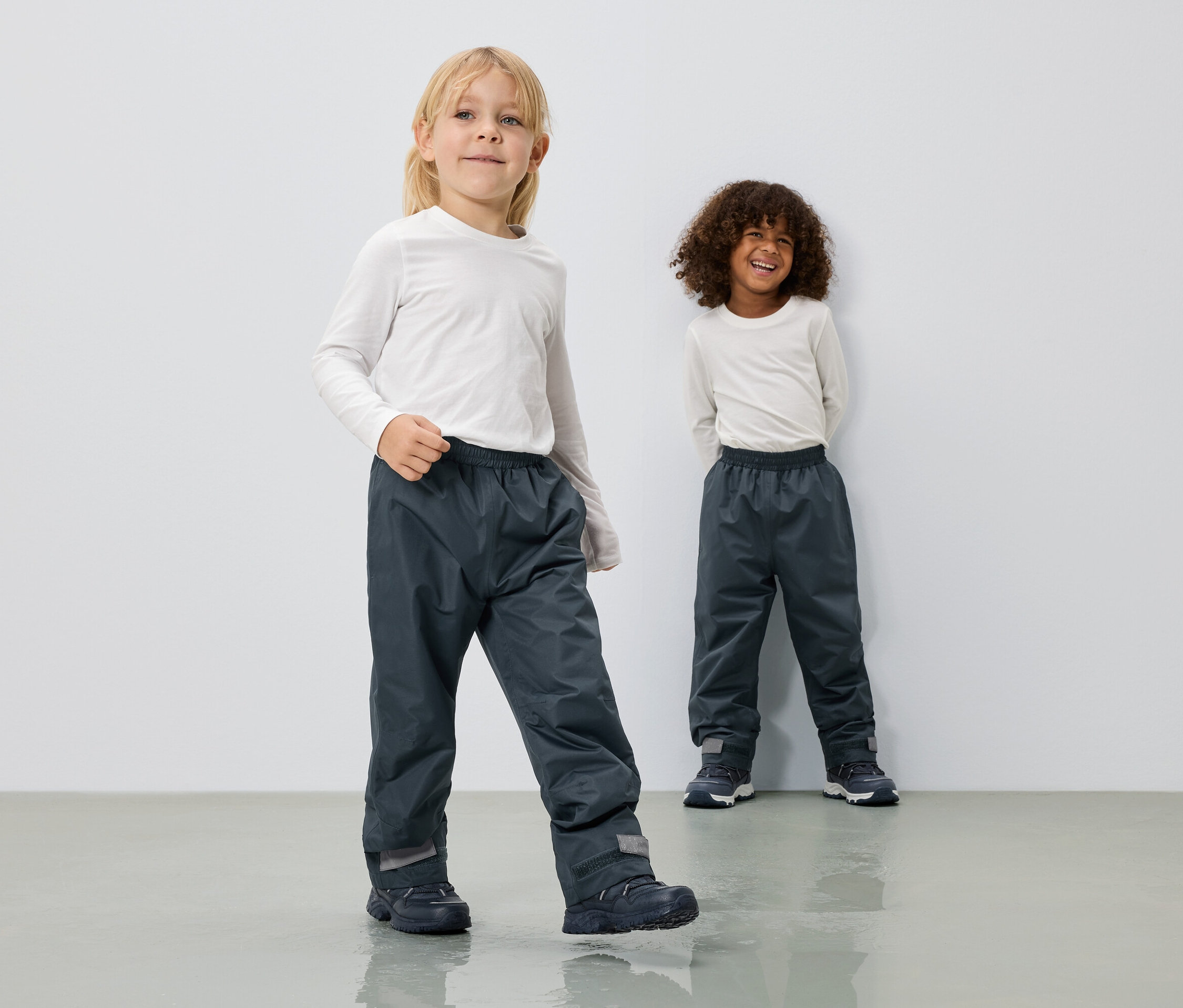 Dve deti majú na sebe dlhé sivé nepremokavé nohavice a biele tričká s dlhým rukávom pred bielou stenou.