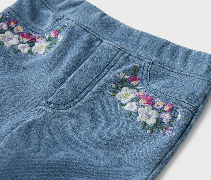 Detailný záber na svetlomodré džínsy s kvetinovou výšivkou na páse a vrecku.