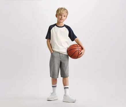 Chlapec s blond vlasmi drží basketbalovú loptu, má na sebe svetlé tričko s tmavomodrými rukávmi, sivé šortky a biele ponožky s čiernymi pruhmi.