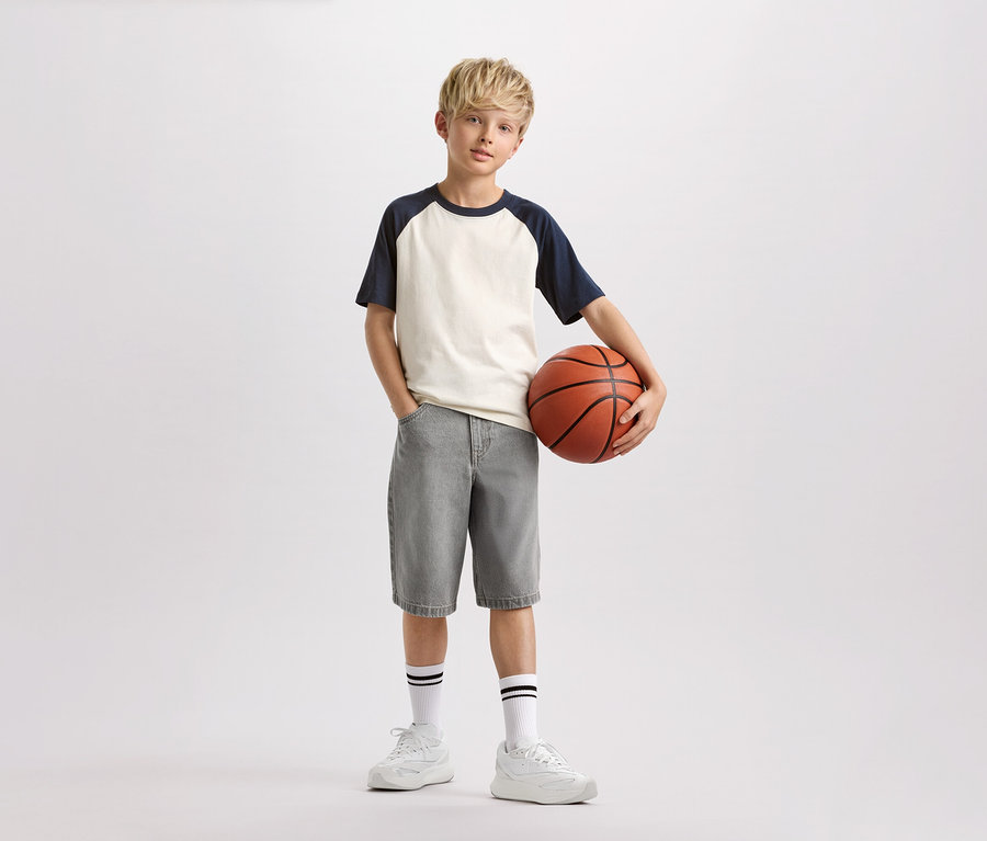 Chlapec s blond vlasmi drží basketbalovú loptu, má na sebe svetlé tričko s tmavomodrými rukávmi, sivé šortky a biele ponožky s čiernymi pruhmi.