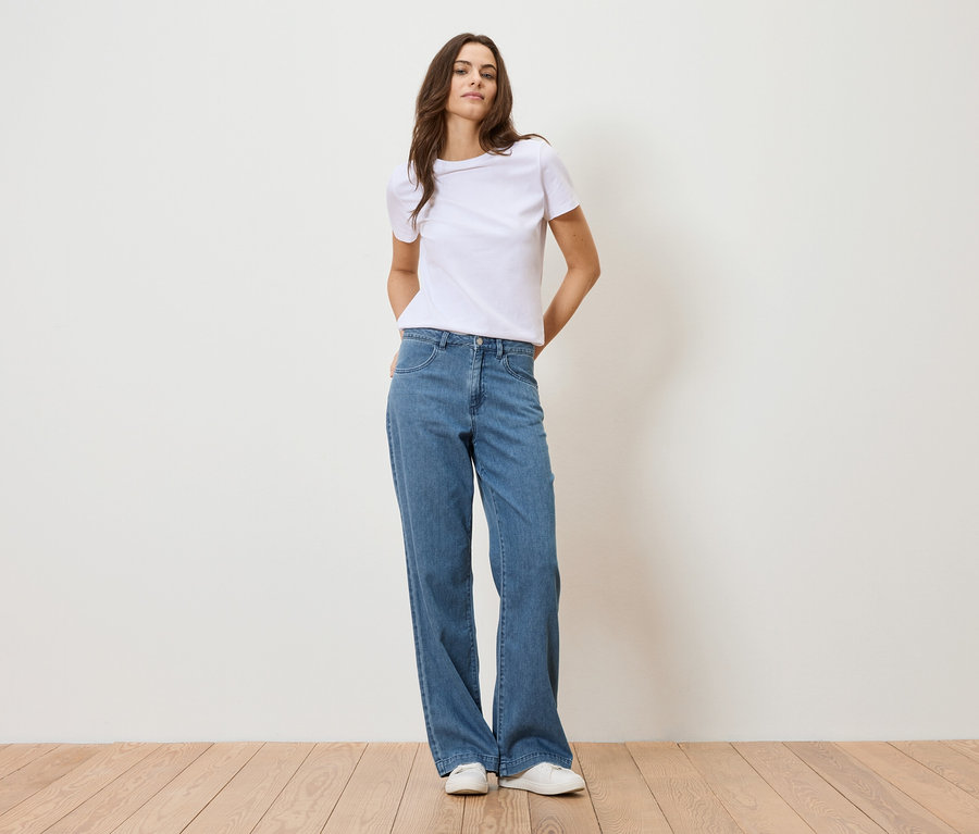 Žena stojí pred bielou stenou a má na sebe biele tričko a modré džínsy.