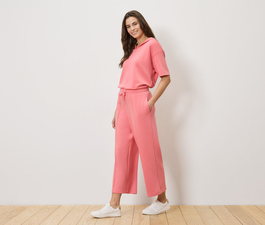 Žena má na sebe koralovú teplákovú súpravu s mikinou s kapucňou a culottes nohavicami a biele tenisky.