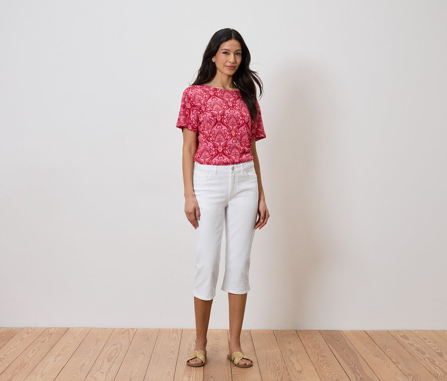 Žena stojí pred stenou a má na sebe červený top a biele nohavice.