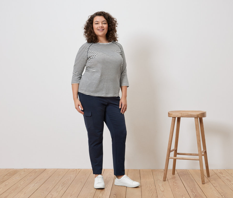 Žena s kučeravými vlasmi stojí v pruhovanom tope, modrých nohaviciach a bielych teniskách vedľa stoličky.