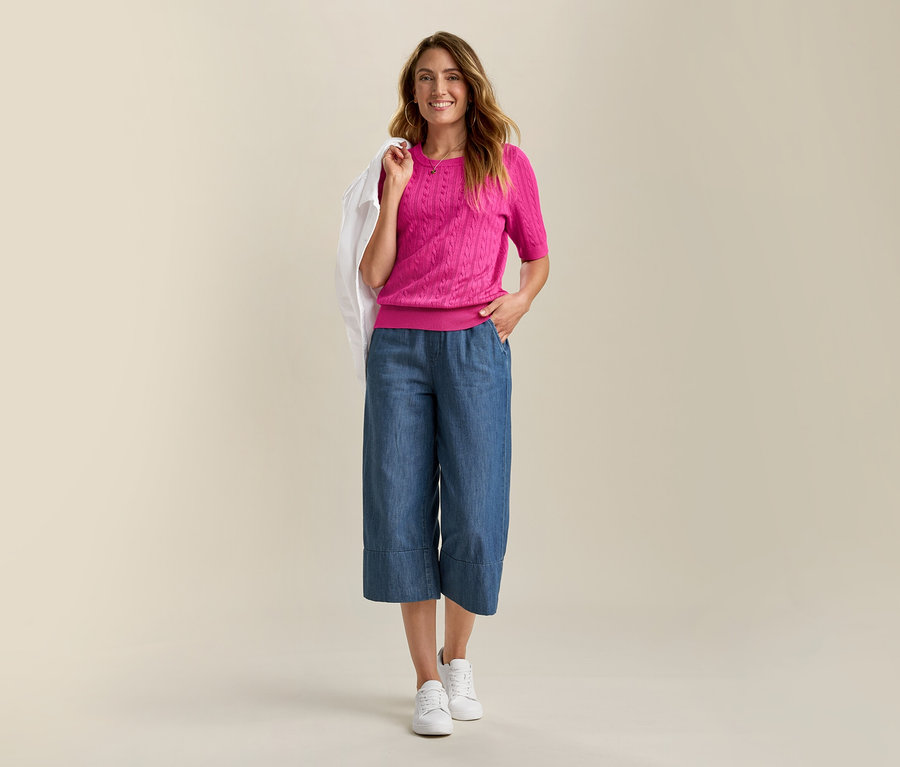 Usmievavá žena má na sebe džínsovú bundu a ružový pulóver s vrkočovým vzorom z jemnej pleteniny. Má na sebe modré culottes a biele tenisky.