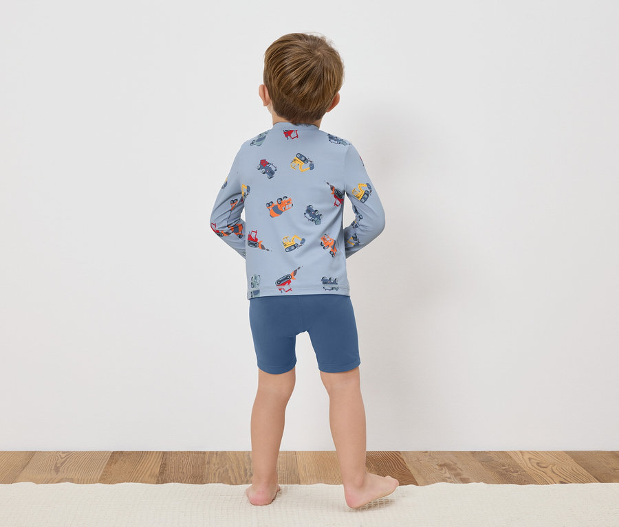 Dieťa stojí chrbtom k fotoaparátu, má na sebe modré tričko s dlhým rukávom s farebnými stavebnými vozidlami a modré šortky.