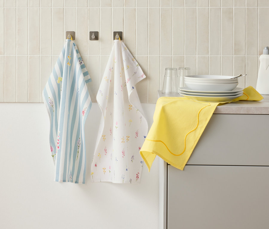 Dve kuchynské utierky visia na hákoch na stene nad kuchynskou linkou, na ktorej je riad, poháre, fľaša na umývanie riadu a žltá handrička.