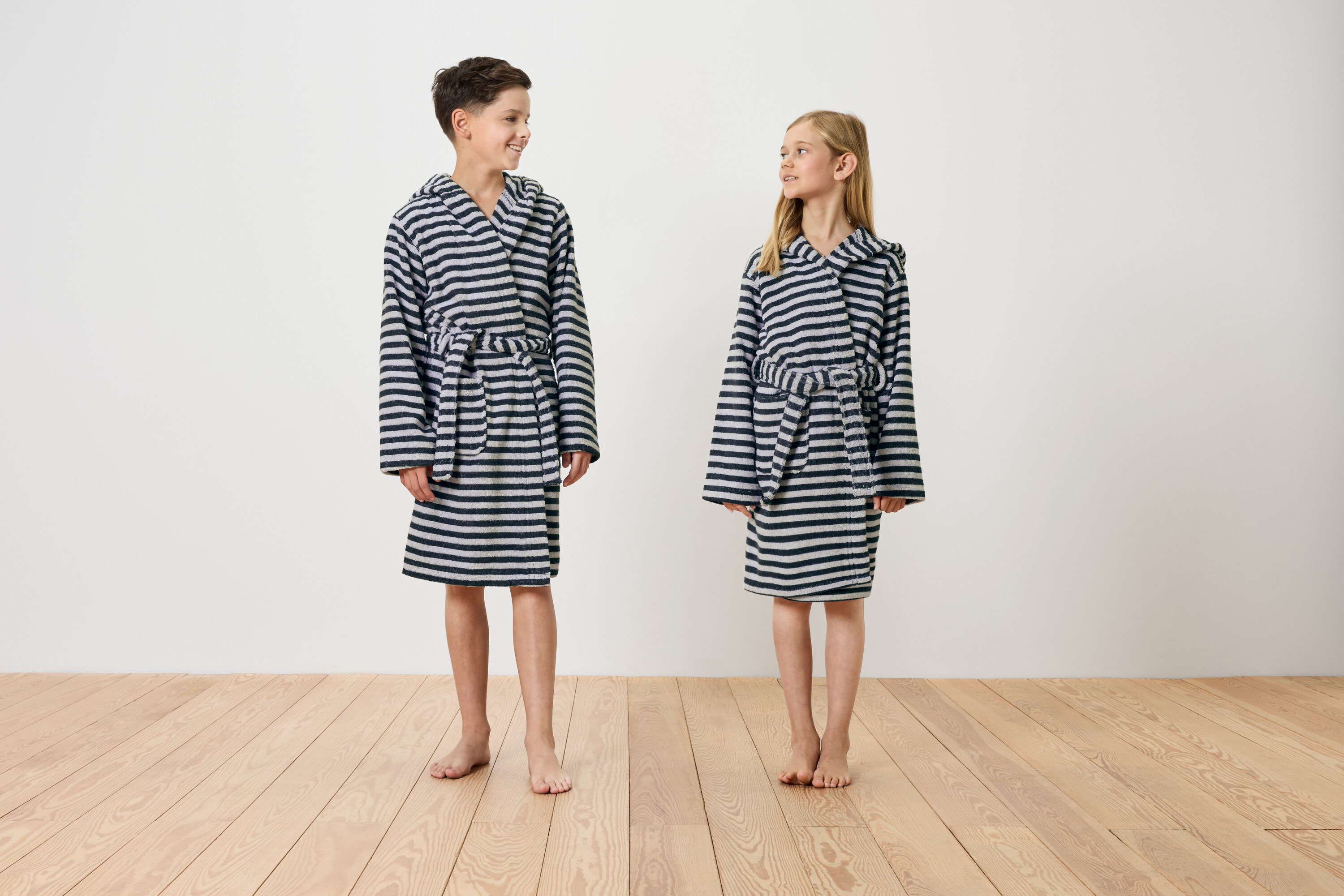 Chlapec a dievča stoja naboso na drevenej podlahe a majú na sebe pruhované župan s kapucňou.