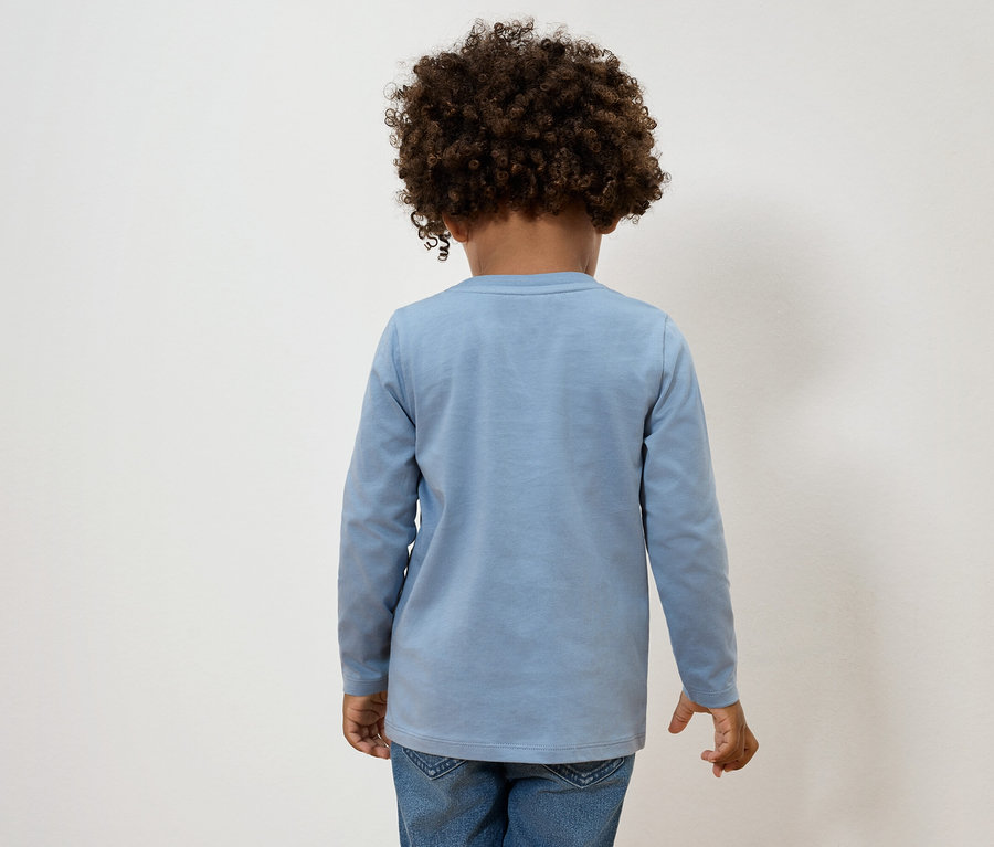 Pohľad zozadu na dieťa s kučeravými vlasmi, oblečené v svetlomodrom tričku s dlhým rukávom a džínsoch.