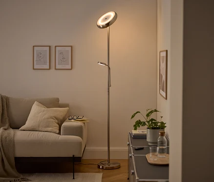 Moderná strieborná stojacia lampa s hlavným svetlom smerujúcim nahor a nastaviteľnou lampou na čítanie osvetľuje útulnú obývačku. Viditeľná je béžová pohovka s dekou a vankúšom, dva zarámované obrazy s linkovou kresbou a tmavá skrinka s rastlinou a fľašou vody.