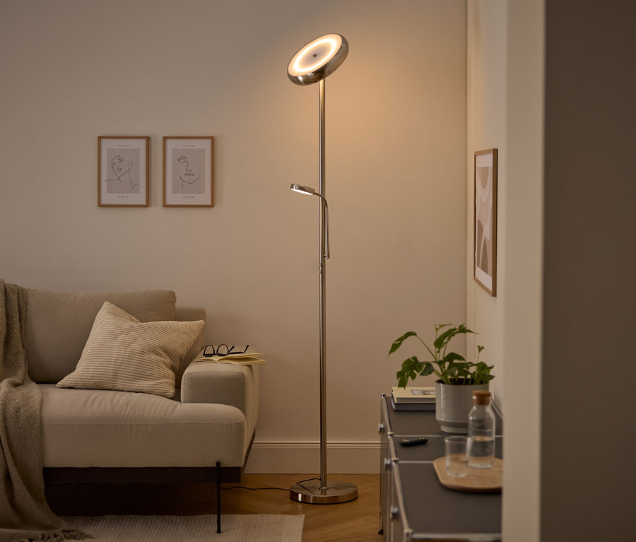 Moderná strieborná stojacia lampa s hlavným svetlom smerujúcim nahor a nastaviteľnou lampou na čítanie osvetľuje útulnú obývačku. Viditeľná je béžová pohovka s dekou a vankúšom, dva zarámované obrazy s linkovou kresbou a tmavá skrinka s rastlinou a fľašou vody.