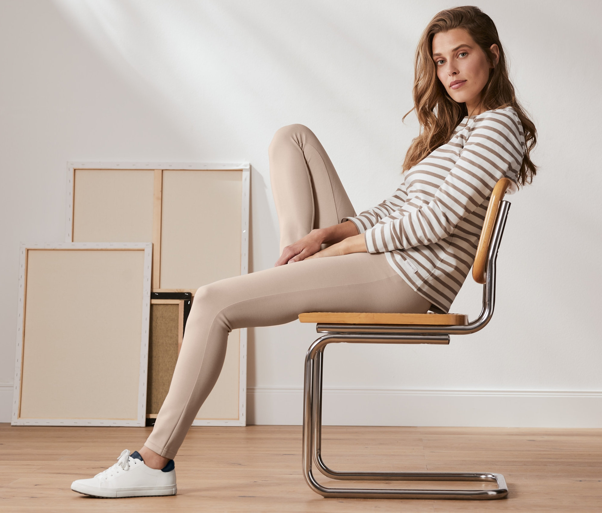 Žena sedí na stoličke a má na sebe krémové jednoduché legínsy z džerseja Heavy Jersey a pásikavú blúzku.