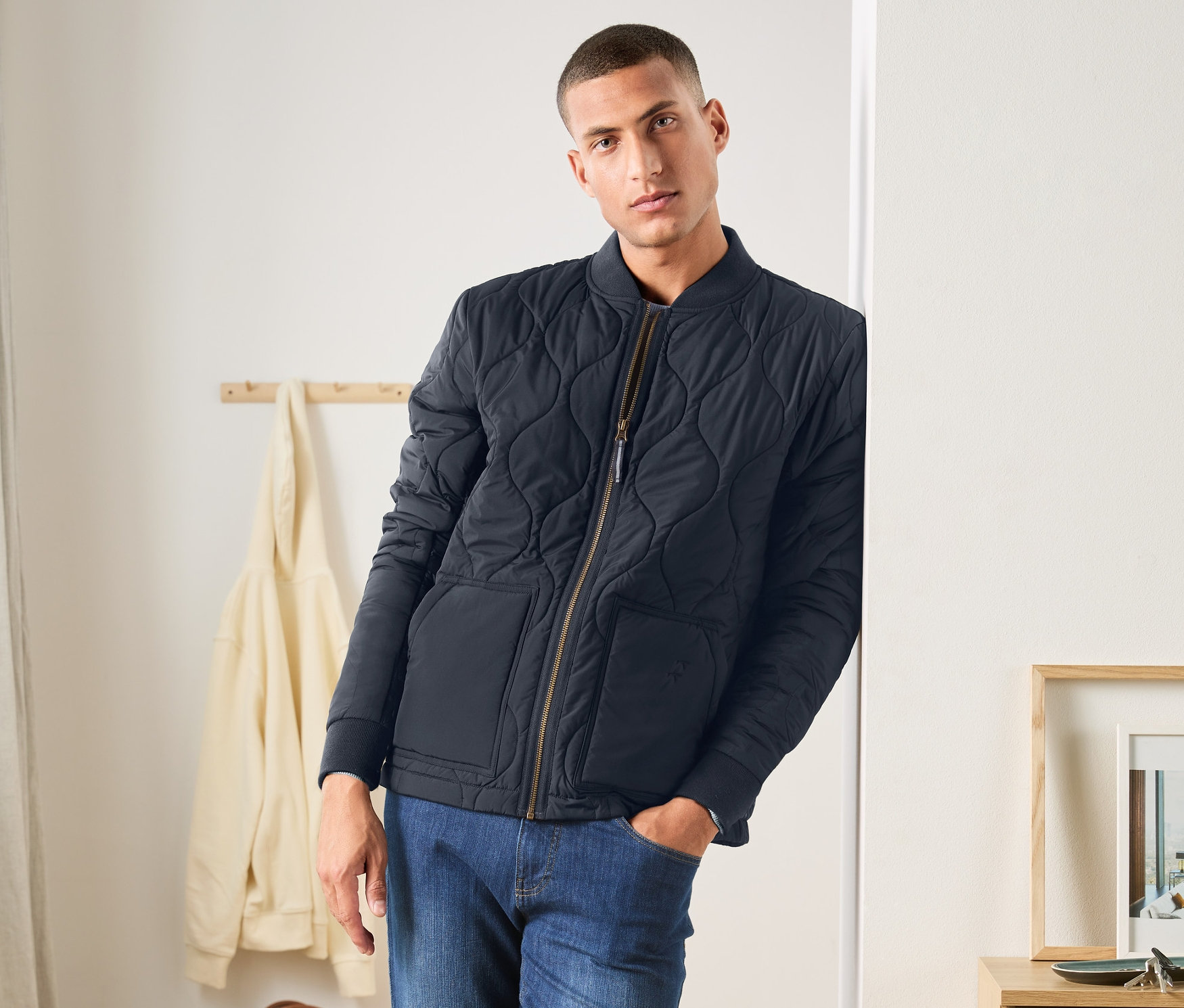 Muž opretý o stenu má na sebe modrú prešívanú bundu, džínsy a biele tenisky.