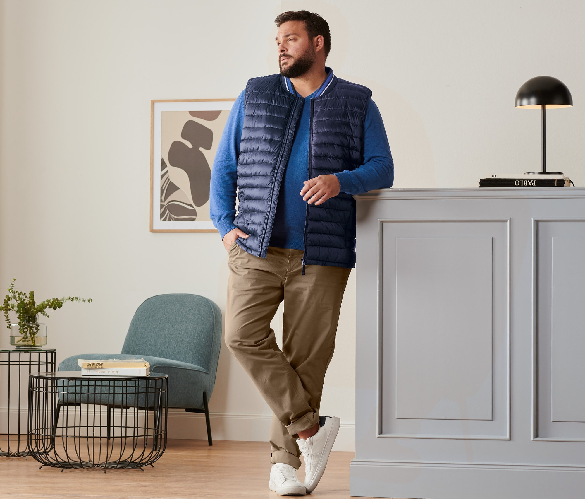 Muž opretý o skrinku má na sebe modrú prešívanú vestu, ľanový pulóver v kráľovskej modrej, nohavice chino vo farbe ťavej srsti a topánky sneaker.