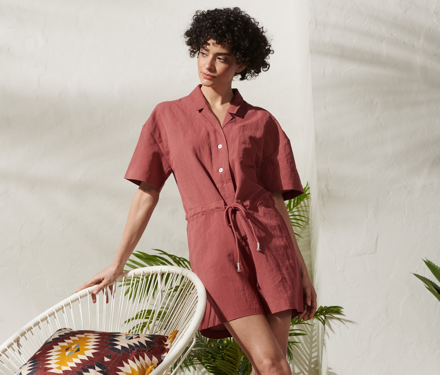 Žena opierajúca sa o biele kreslo v krátkom overale z ľanovej zmesi.