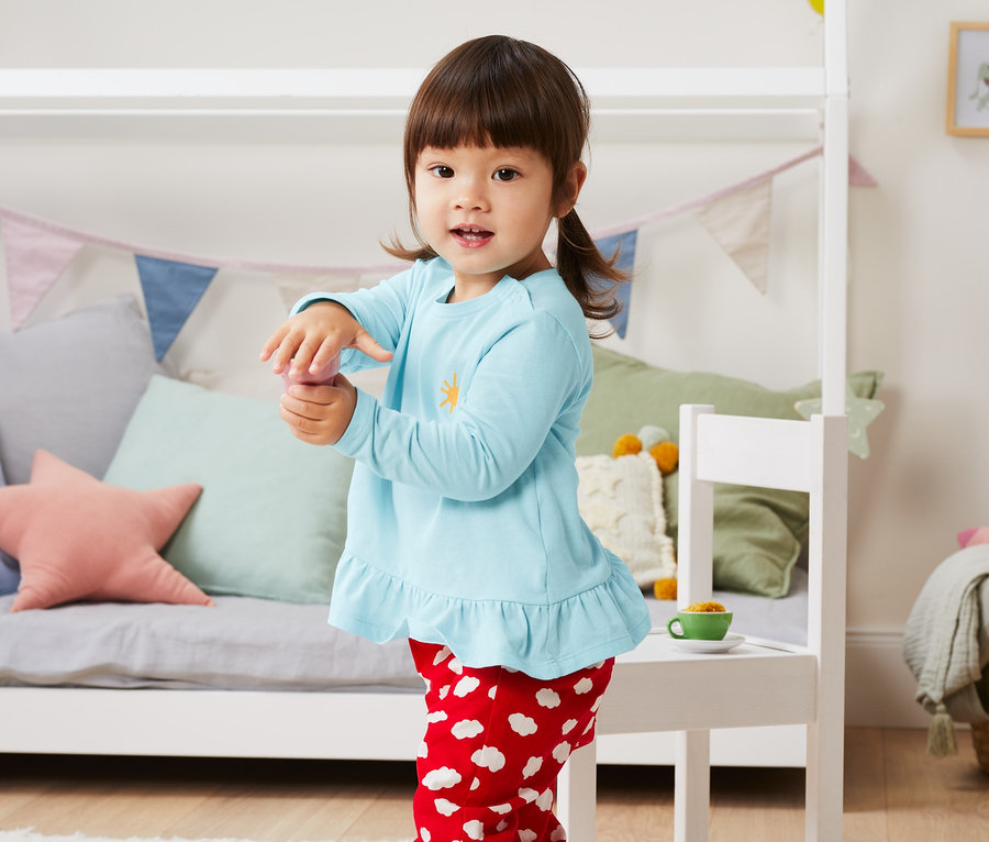 Dievča stojí v detskej izbe a má na sebe svetlomodré tričko a červené nohavice s bielymi obláčikmi. Na sebe má tričká z biobavlny, 3 ks.