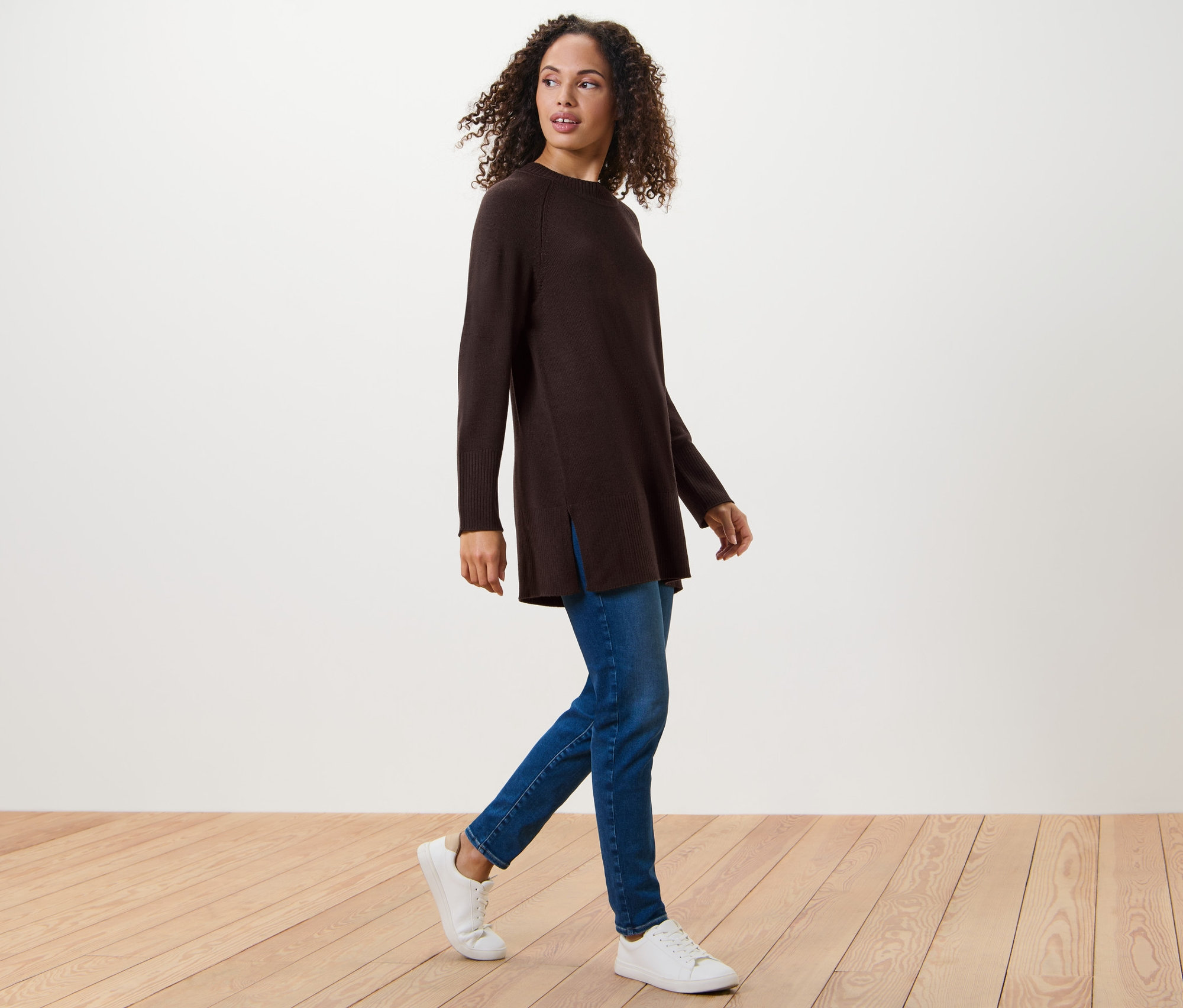 Žena kráča a má na sebe dlhý pletený pulóver tmavohnedej farby, džínsy a tenisky.