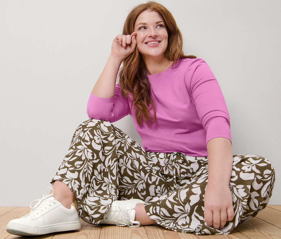 Žena sedí na zemi v ružovom tričku s trojštvrťovými rukávmi a nohaviciach s potlačou.