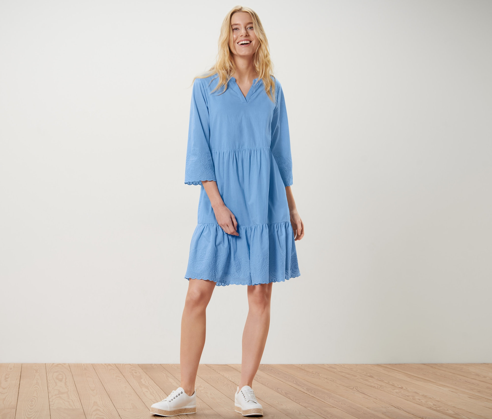 Blondínka stojí a usmieva sa, má na sebe modré tkané šaty s výšivkou a biele tenisky.