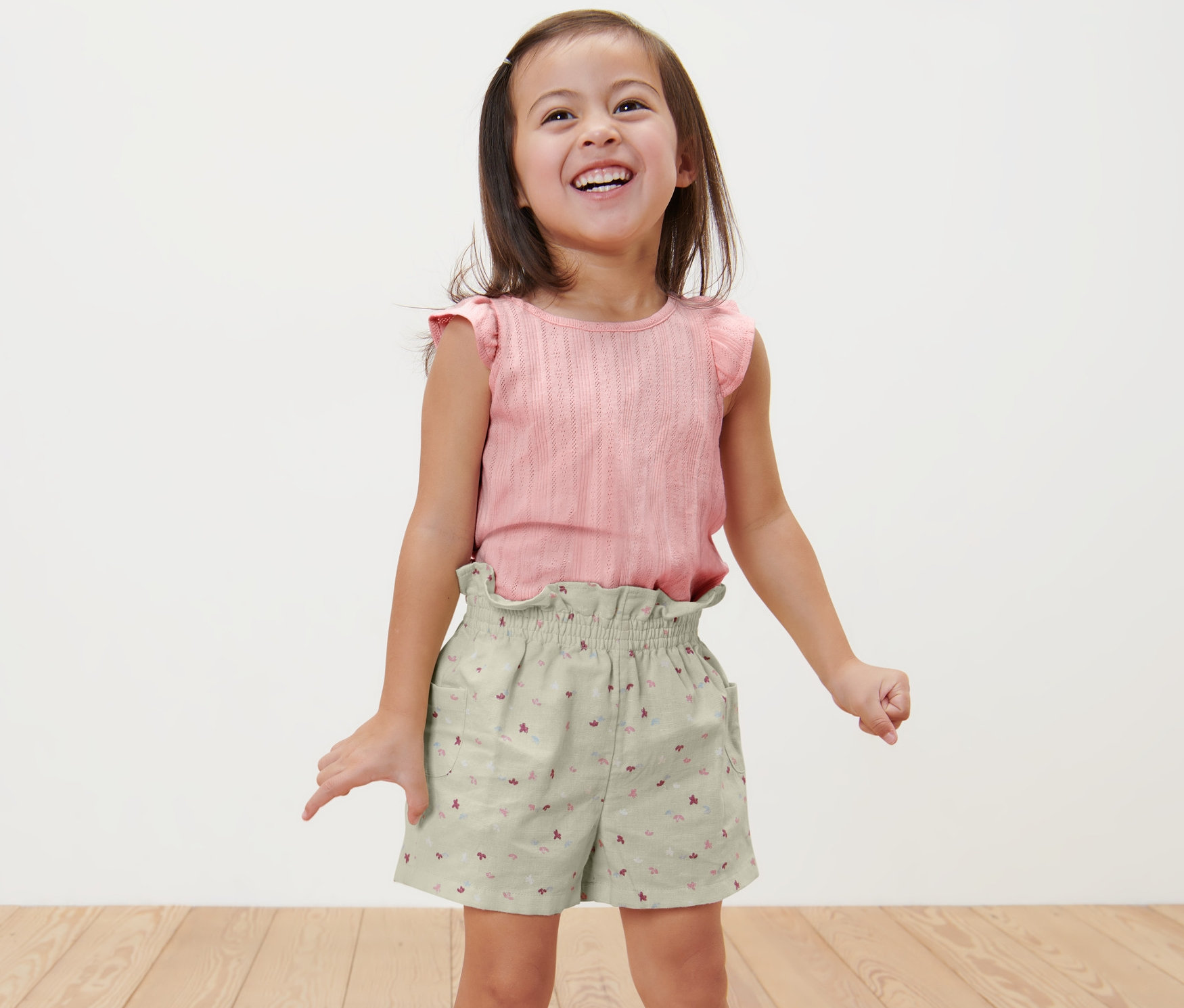 Usmievajúce sa dievčatko stojí a má na sebe detské šortky v štýle paper bag a ružový top.