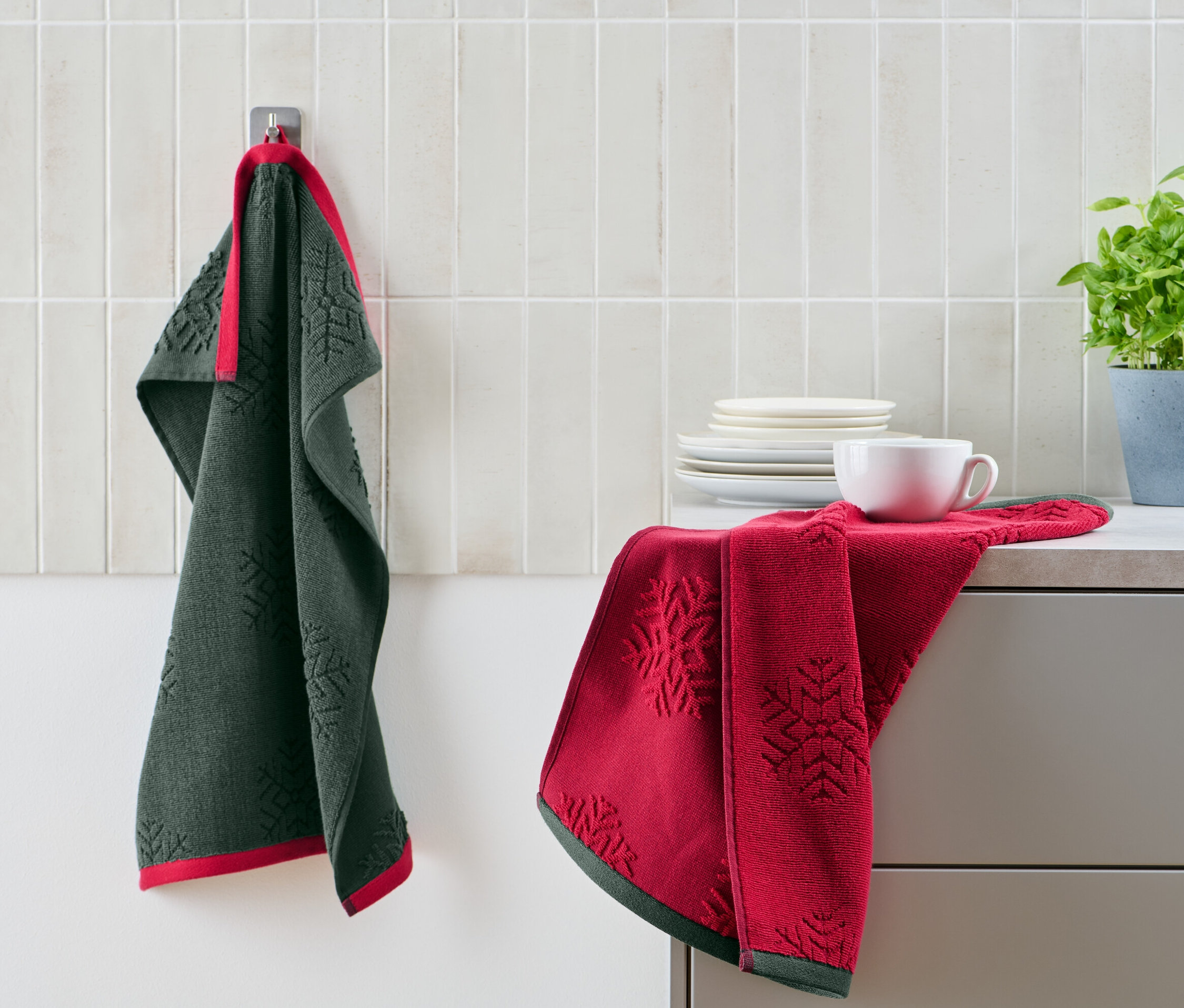 Zelená kuchynská utierka visí na háčiku vedľa červenej utierky a šálky na pulte. V pozadí sú taniere a rastlina v kvetináči.