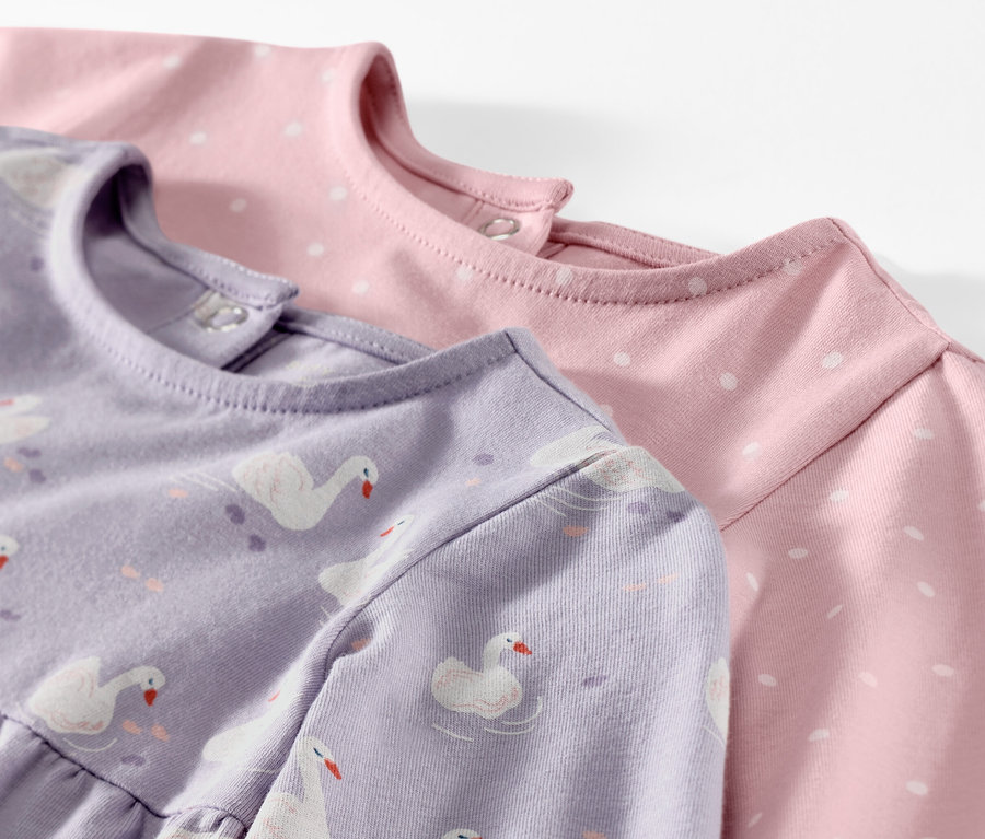 Detail dvoch dojčenských džersejových šiat, jedny fialové so vzorom labutí, druhé ružové s bielymi bodkami.