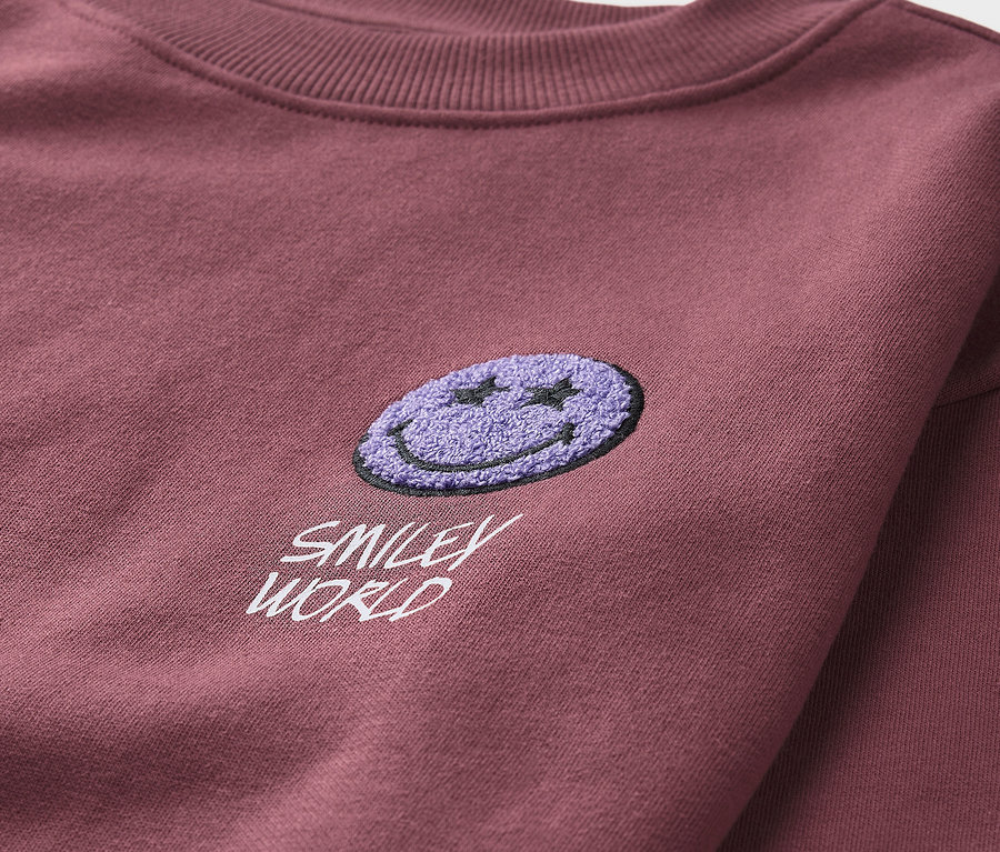 Detail bordovej detskej mikiny SMILEYWORLD® s nášivkou fialového smajlíka.