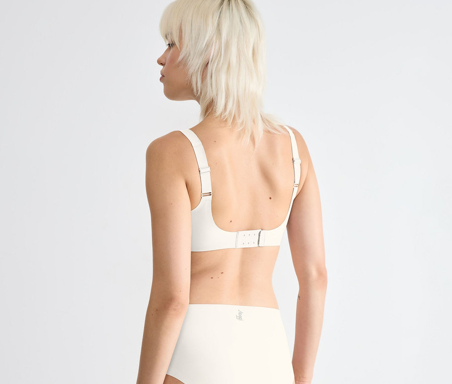 Žena s krátkymi blond vlasmi, videná zozadu, má na sebe sloggi ZERO Feel 2.0 Soft Bra vo farbe silk white a zladené nohavičky s vysokým pásom.