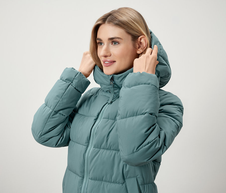 Žena si upravuje kapucňu na tyrkysovej páperovej bunde.